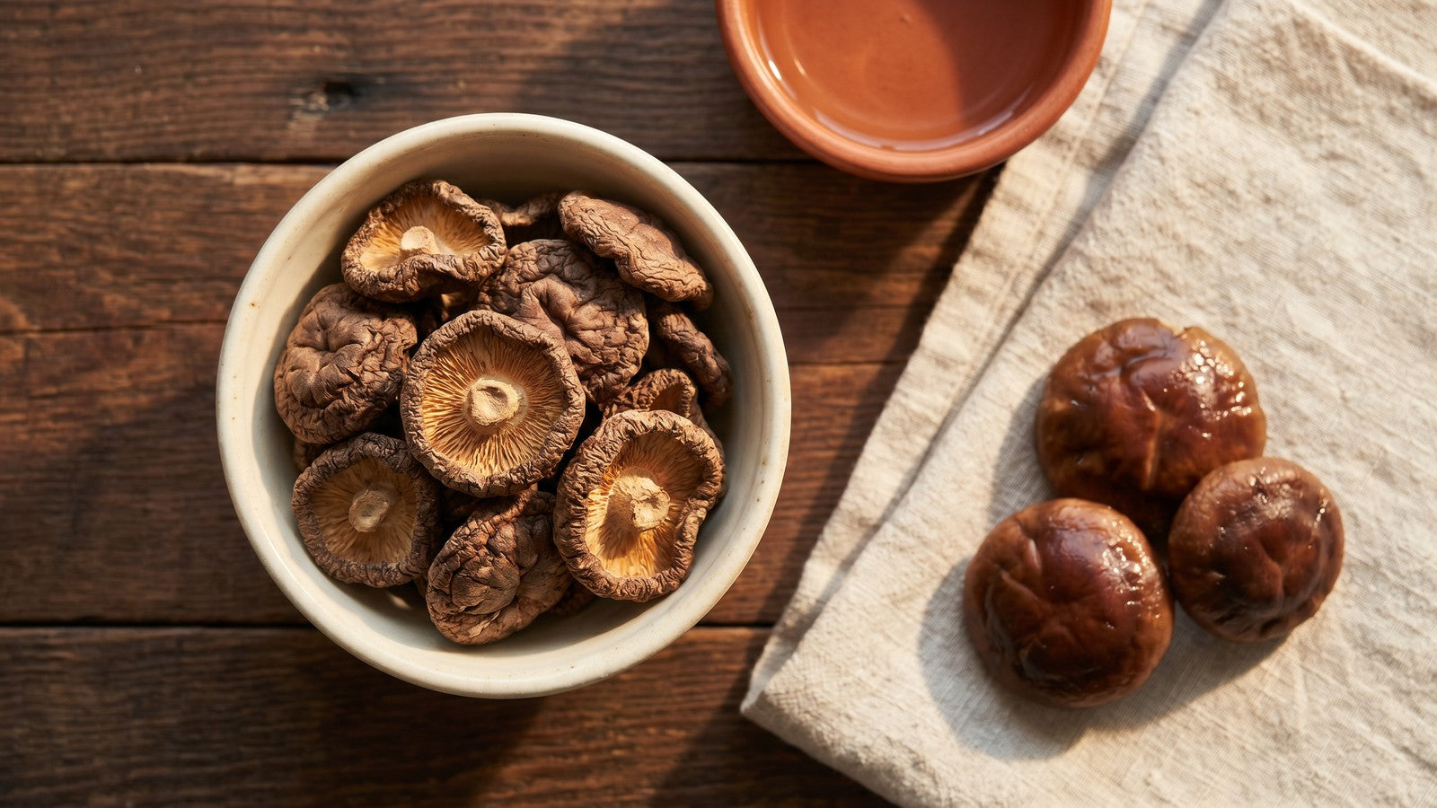 Dried shiitake mushrooms in a ceramic bowl beside a few rehydrated ones on a rustic wood surface