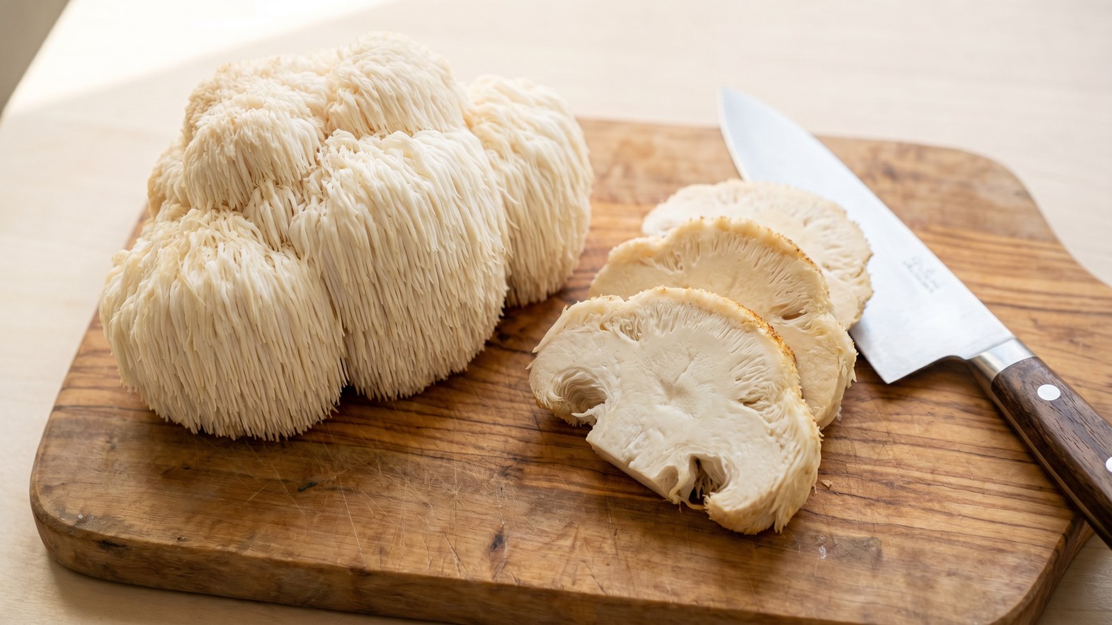 Fresh whole lion's mane mushroom cluster on a wood cutting board with sliced pieces showing the pom-pom texture